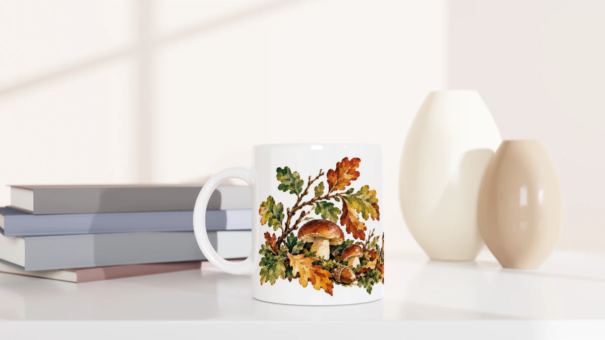 Weiße Tasse mit herbstlicher Waldszene aus Igel, Laub, Pilzen und Eicheln in Ocker-, Braun-, Grün- und Rosttönen.