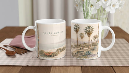 Seitliche Ansicht der Santa Monica California Tasse mit aquarelliertem Küstenweg, Holzgeländer, Pazifik, hellem Strand, Bergen und einer Parkbank unter kalifornischen Palmen